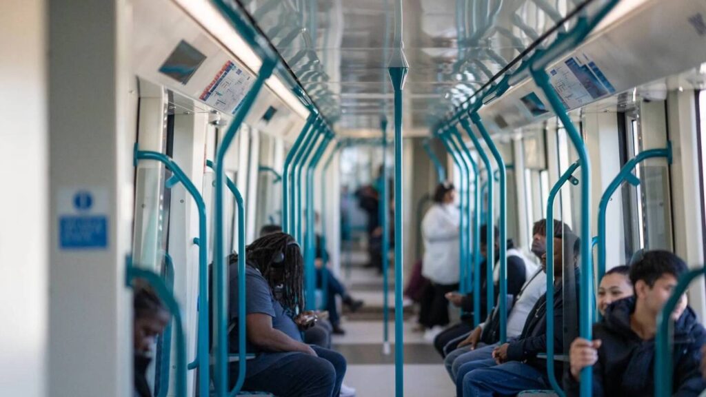 Passengers sitting inside a new walk-through DLR train with turquoise handrails and large windows, showing a bright and open carriage design.