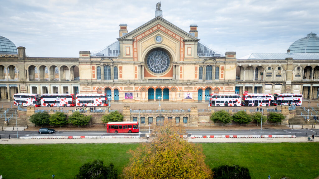 Several red double decker London buses decorated with poppy graphics parked outside Alexandra Palace for London Poppy Day.