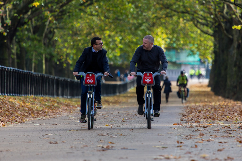 Two men riding Santander Cycles through a tree lined path in autumn